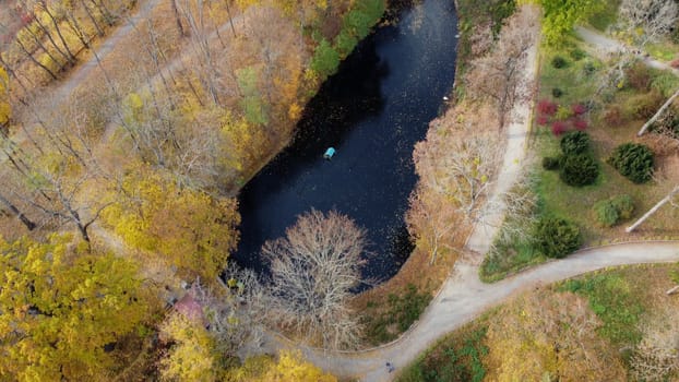 Flying over the autumn park. Many trees with yellow green and fallen leaves, lakes, people walking along dirt paths in park on autumn day. Top view. Aerial drone view. Beautiful natural background