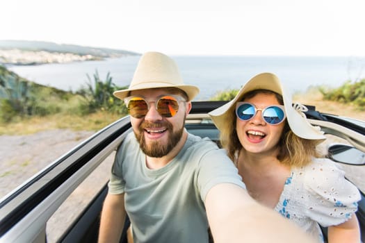 Happy beautiful couple in love taking a selfie portrait driving a convertible car on the road at vacation. Rental cars and vacation concept