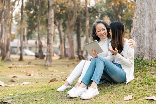 Lovely granddaughter and grandmother sitting under the tree in the green park together, watching video on tablet, sipping coffee.