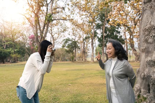 Lovely Asian granddaughter taking a picture of her happy grandmother with camera while strolling in the green park together.
