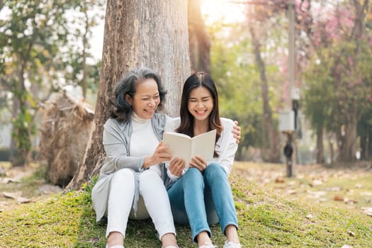Lovely granddaughter and grandmother sitting under the tree in the green park, read a book together.