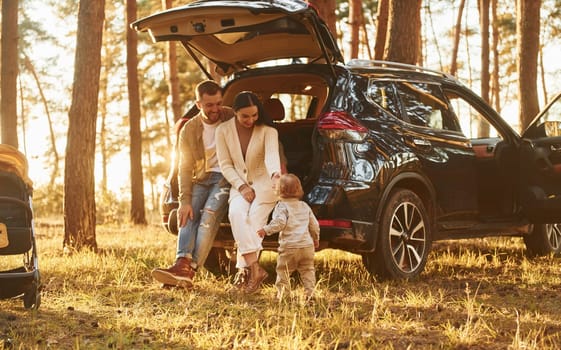 Leaning on the car. Happy family of father, mother and little daughter is in the forest.