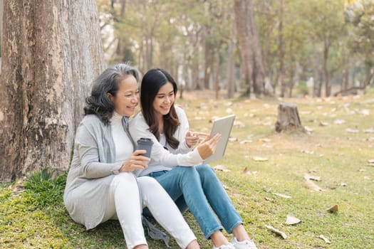 Lovely granddaughter and grandmother sitting under the tree in the green park together, watching video on tablet, sipping coffee.
