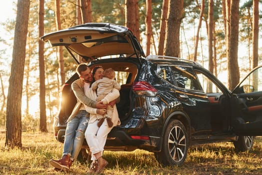 Leaning on the car. Happy family of father, mother and little daughter is in the forest.