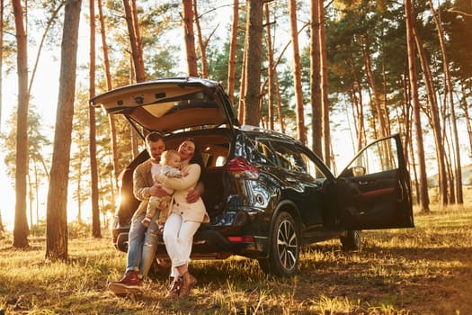 Leaning on the car. Happy family of father, mother and little daughter is in the forest.
