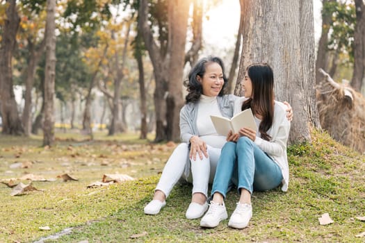 Lovely granddaughter and grandmother sitting under the tree in the green park, read a book together.