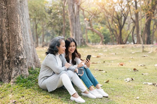 Lovely granddaughter and grandmother sitting under the tree in the green park together, watching Social news on telephone, sipping coffee.