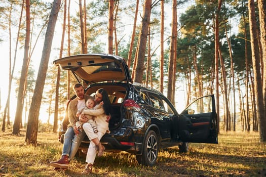 With black colored automobile. Happy family of father, mother and little daughter is in the forest.
