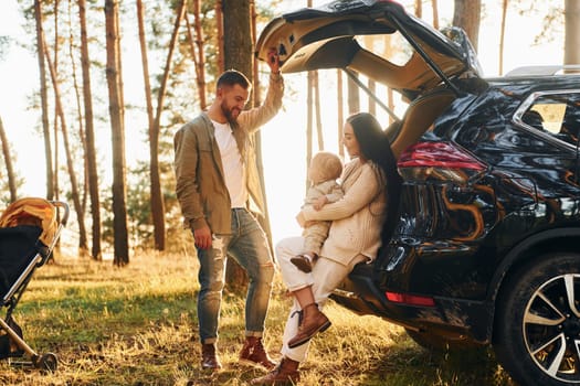 With black colored automobile. Happy family of father, mother and little daughter is in the forest.