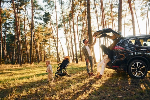 With black colored automobile. Happy family of father, mother and little daughter is in the forest.