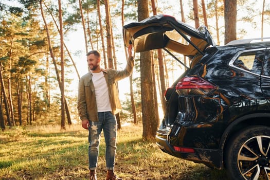 Near the car. Man in jeans is outdoors in the forest with his black colored automobile.