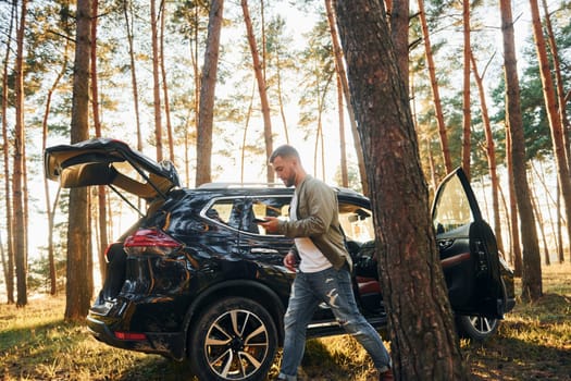 Near the car. Man in jeans is outdoors in the forest with his black colored automobile.