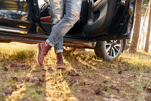 View of jeans and shoes. Man in jeans is outdoors in the forest with his black colored automobile.
