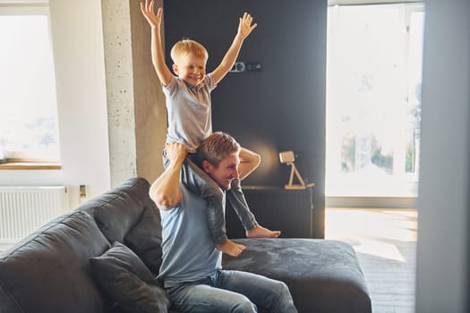 Boy is sitting on the man's shoulders. Father and son is indoors at home together.