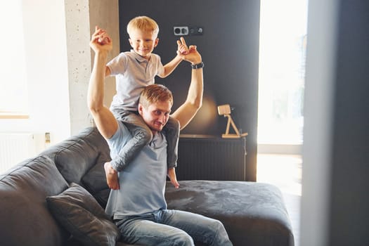 Boy is sitting on the man's shoulders. Father and son is indoors at home together.