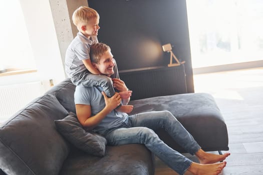 Boy is sitting on the man's shoulders. Father and son is indoors at home together.