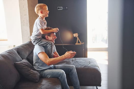 Boy is sitting on the man's shoulders. Father and son is indoors at home together.