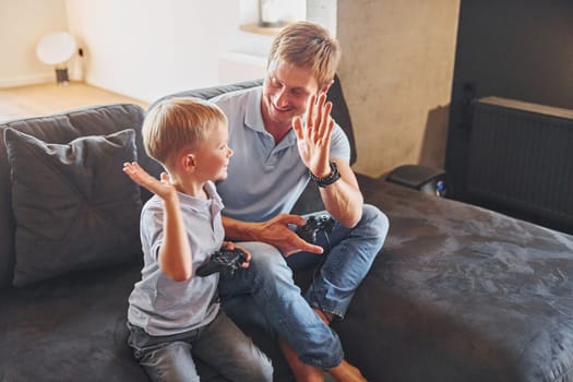 Playing video game. Father and son is indoors at home together.