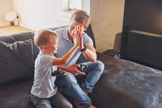 With video game joysticks. Father and son is indoors at home together.