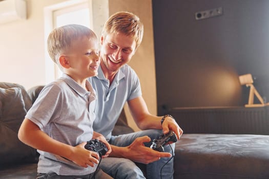 With video game joysticks. Father and son is indoors at home together.
