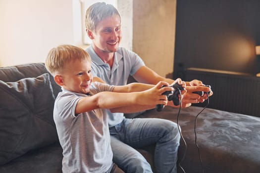 With video game joysticks. Father and son is indoors at home together.