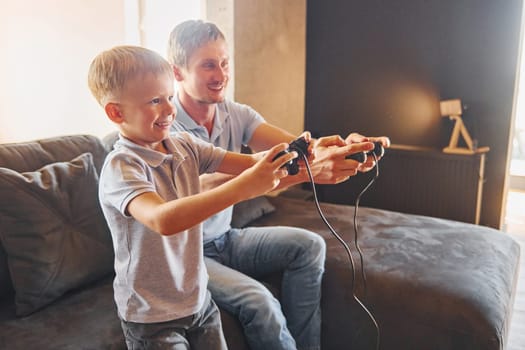With video game joysticks. Father and son is indoors at home together.