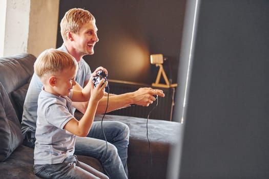 Holding video game controllers. Father and son is indoors at home together.