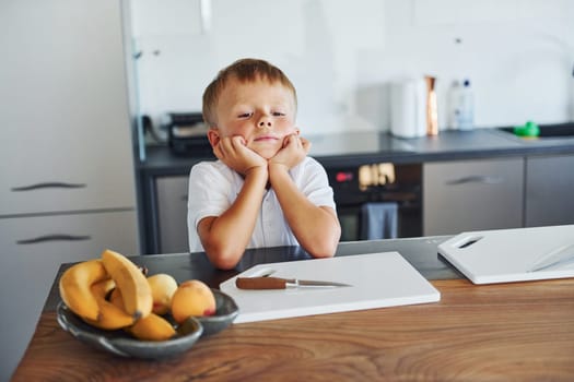 Boy in casual clothes is sitting in the kitchen with bananas on table.