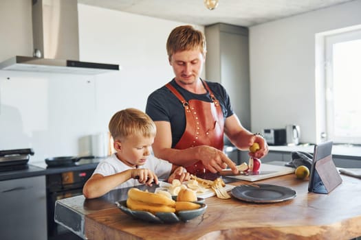 Man is in the apron on kitchen. Father and son is indoors at home together.