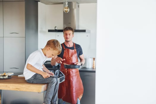 Man is in the apron on kitchen. Father and son is indoors at home together.