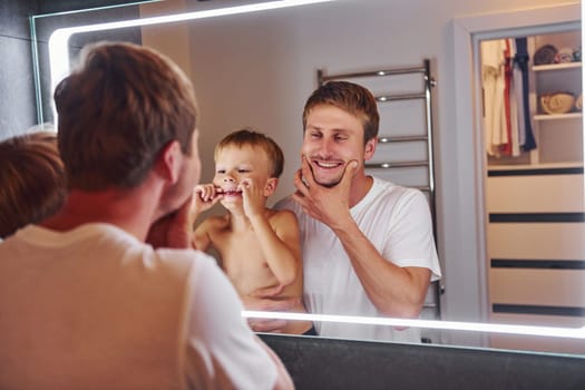 Looking in the mirror in bathroom. Father and son is indoors at home together.
