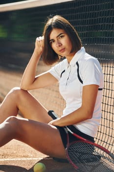 Sitting and holding racket. Female tennis player is on the court at daytime.