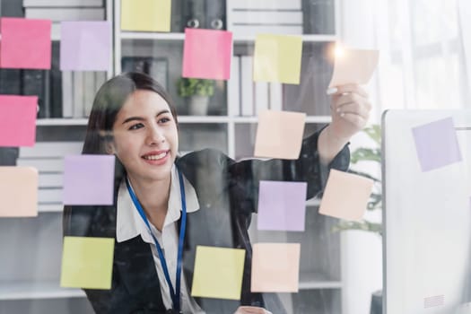 Young businesswoman creative team using post it notes in glass wall to writing strategy business plan to development grow to success...