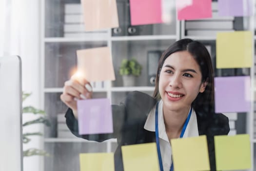 Young businesswoman creative team using post it notes in glass wall to writing strategy business plan to development grow to success...