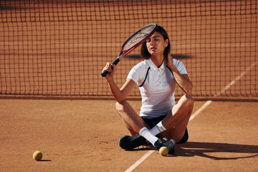 Brunette is sitting on the ground. Female tennis player is on the court at daytime.