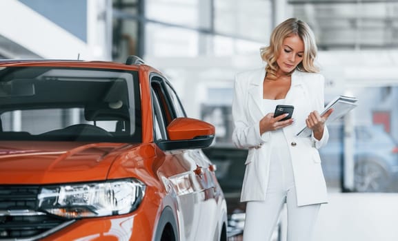 Holding phone. Woman in formal clothes is indoors in the autosalon.