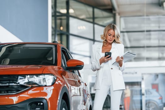 Holding phone. Woman in formal clothes is indoors in the autosalon.