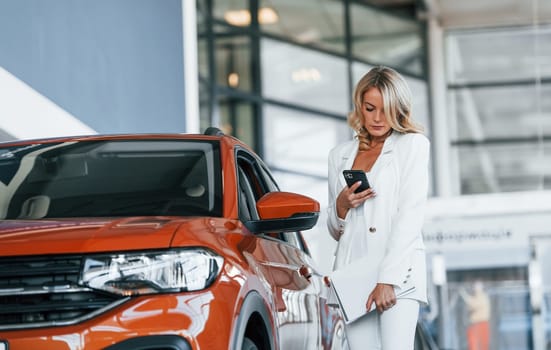 Holding phone. Woman in formal clothes is indoors in the autosalon.
