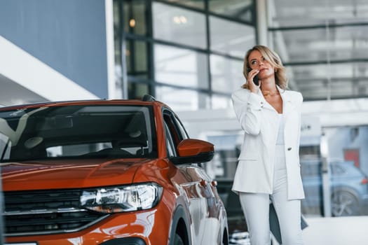 Holding phone. Woman in formal clothes is indoors in the autosalon.