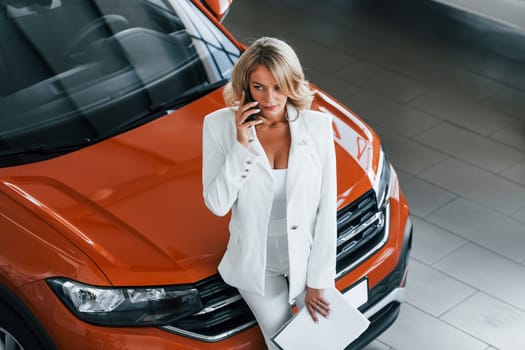 Holding phone. Woman in formal clothes is indoors in the autosalon.