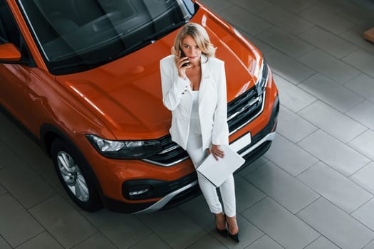 With phone in hand. Woman in formal clothes is indoors in the autosalon.