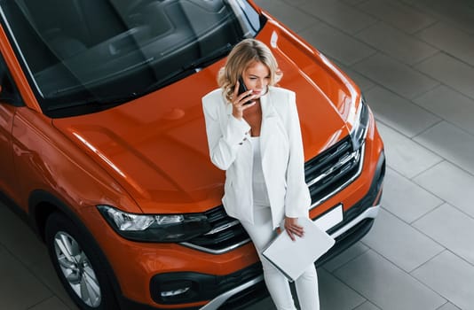 With phone in hand. Woman in formal clothes is indoors in the autosalon.