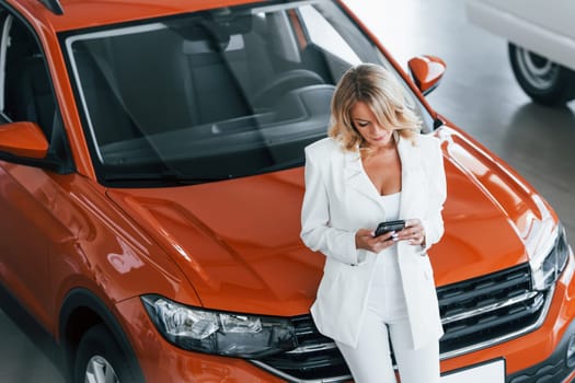 With phone in hand. Woman in formal clothes is indoors in the autosalon.