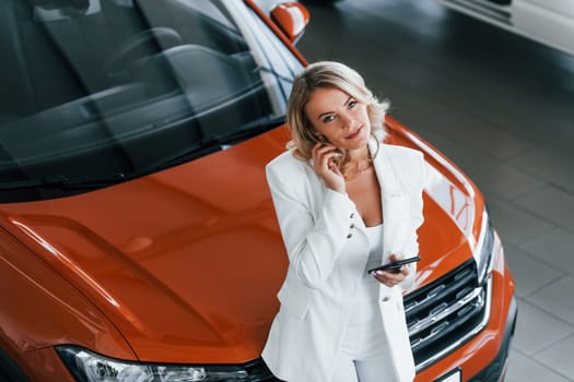 Leaning on the car. Woman in formal clothes is indoors in the autosalon.