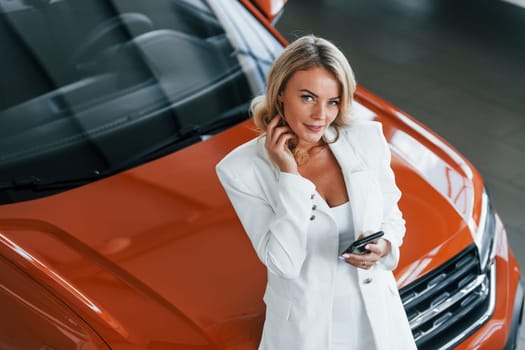 Leaning on the car. Woman in formal clothes is indoors in the autosalon.
