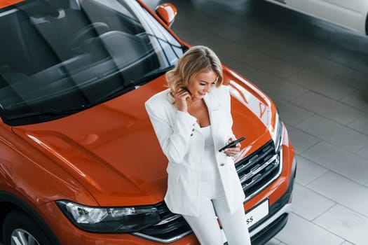 Leaning on the car. Woman in formal clothes is indoors in the autosalon.