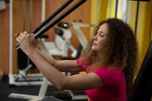 Performing weight training exercises. Lifting weights at a gym. A well-equipped exercise room. Workout performed at the gym. A woman uses sports equipment to exercise her upper body.