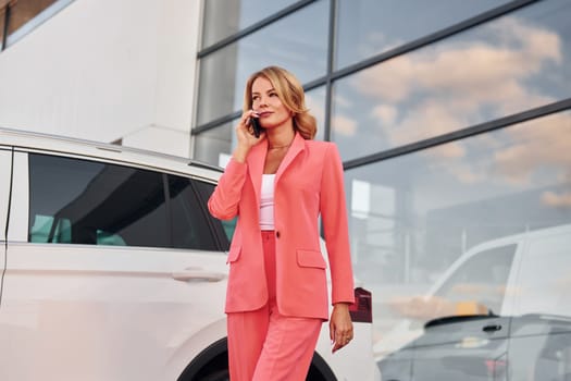 Having conversation by phone. Woman in formal wear standing outdoors with phone in hands and near car.