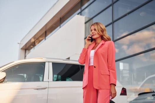 Having conversation by phone. Woman in formal wear standing outdoors with phone in hands and near car.