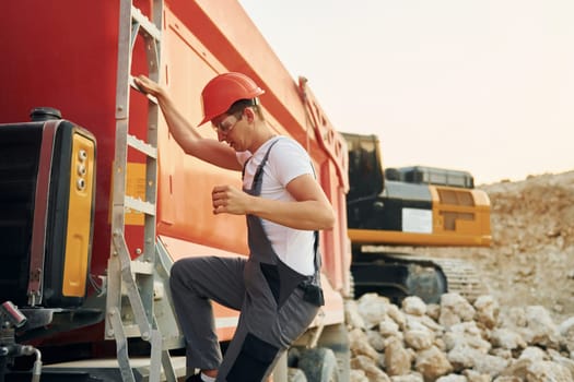 Going up to the vehicle seat. Worker in professional uniform is on the borrow pit at daytime.